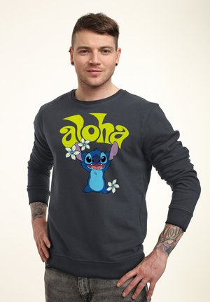 Mand med kort hår og tatoveringer iført en mørk sweatshirt med Stitch-figuren, "aloha"-tekst og hvide blomster.