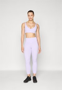 Reggiseno sportivo lilla con un design liscio e senza cuciture e spalline larghe, abbinato a leggings a vita alta coordinati, completato da scarpe da ginnastica nere.
