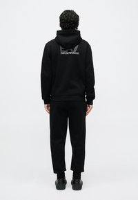 Άνδρας στέκε�ται με την πλάτη προς τα εμπρός, φορώντας μαύρη φούτερ με κουκούλα EA7 Emporio Armani, μαύρο παντελόνι που ταιριάζει και μαύρα αθλητικά παπούτσια, σε λευκό φόντο.