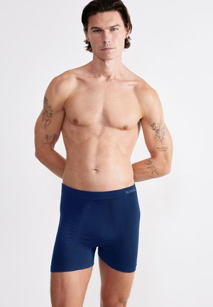 Skjorteløs mand med tatoveringer står vendt fremad iført marineblå boxershorts mod en ensfarvet hvid baggrund.