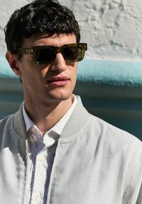 Gafas de sol de montura cuadrada en plástico verde oliva con lentes oscuros, combinadas con una chaqueta bomber gris claro sobre una camisa blanca con estampado.
