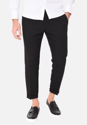 Uomo che indossa pantaloni neri aderenti corti, camicia bianca e sneakers slip-on nere con suole bianche, in piedi con le mani nelle tasche.