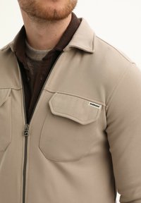 Hombre que lleva una chaqueta beige con cremallera y bolsillos con solapa sobre una camisa marrón abotonada y un suéter gris de punto visible en el cuello.