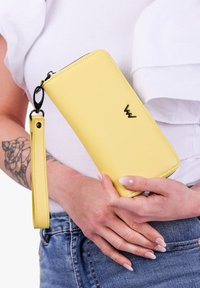 Yellow leather wallet with a black logo, zip closure, detachable wrist strap, and smooth texture, held against a white top.