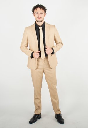 Costume beige ajusté, à revers crantés, avec une cravate assortie sur une chemise noire. Des chaussures de ville noires complètent la tenue.