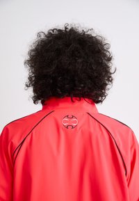 Red waterproof jacket featuring a black logo on the upper back, with black piping along the seams and a smooth, lightweight texture.