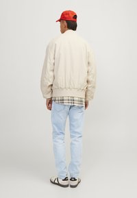 Beige bomberjack met geribbelde zoom, laag over een geruite shirt, gecombineerd met lichtblauwe spijkerbroek en zwart-witte sneakers. Rode pet.