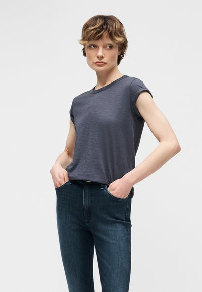 Νεαρός που φοράει γκρι σκουρόχρωμο T-shirt με γυρισμένα μανίκια και σκούρο μπλε τζιν, στέκεται με τα χέρια στις τσέπες, κοιτάζοντας προς τα πλάγια.