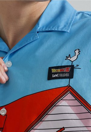 Chemise à manches courtes, de couleur bleu clair, présentant un graphique de maison rouge et blanc, ainsi qu'un écusson noir DRAGON BALL Z cousu sur la poitrine.