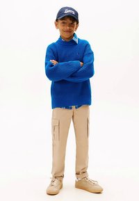 Niebieski sweter z dzianiny na niebieskiej koszuli w kratę, beżowe spodnie cargo z bocznymi kieszeniami i jasnobrązowe trampki. Ciemna czapka z białymi akcentami logo.