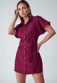 Lühikeste varrukatega nööpidega kleit burgundia värvi kangast, kahel eesmisel taskul ja sobiva vööga. Omab struktureeritud kraed ja sujuvat tekstuuri.