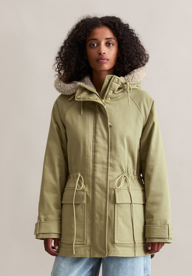 Khaki groene parka jas met een met bont gevoerde capuchon, een koord in de taille en twee voorklauwen. Heeft gouden hardware en een stevige buitenstof.