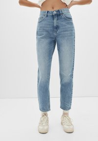Jeans taille haute en denim bleu clair avec une coupe slim, présentant des zones décolorées et un design classique à cinq poches, associés à des baskets beiges.
