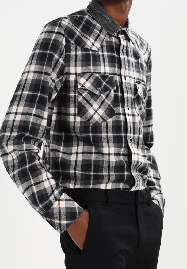 Personne portant une chemise à carreaux noir et blanc à manches longues avec des poches poitrine boutonnées et un pantalon noir, debout les mains dans les poches.