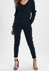 Navyblauwe gebreide trui met V-hals en gerafelde mouwen, gecombineerd met slim-fit broek met een tweeknoopsluiting en zijstrepen.