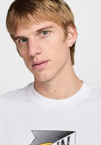 Camiseta blanca con un diseño gráfico en negro y amarillo en la parte frontal. El modelo tiene el cabello corto y rubio, y múltiples piercings en la oreja.