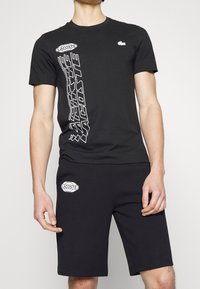 Homme portant un t-shirt noir Lacoste avec un logo vertical et un short noir assorti avec un écusson Lacoste sur la jambe gauche.