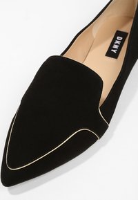 Chaussure plate noire à bout pointu avec un détail de bordure dorée et un intérieur beige, avec une étiquette DKNY à l'intérieur.