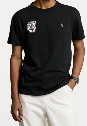 Mand iført en sort t-shirt med et Tysklandslogo på venstre bryst og et lille gult polospiller-logo på højre bryst, kombineret med hvide bukser.