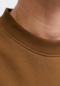 Nærbillede af en persons nakke iført en brun ribbet crewneck-sweater med synlig hudtekstur.