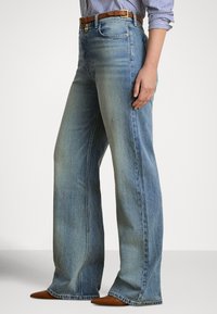 Sieviete, kas valkā gaiši zilas platām kājām džinsas, brūnas smailas kurpes, brūnu jostu un zilu svītrainu krekli ar uzrullētām piedurknēm.