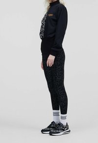 Sweatshirt de sport noir avec fermeture éclair sur le côté, ayant un motif géométrique et un logo ; associé à un legging noir et des baskets à motifs. Tissu texturé.