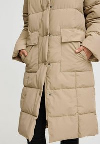 Abrigo acolchado beige con un diseño acolchado, que presenta dos bolsillos frontales, botones a presión y un cierre con cremallera en la parte inferior. Textura suave y corte relajado.