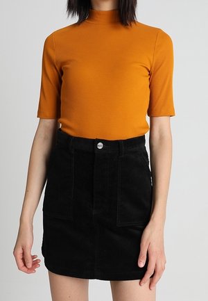 Kobieta ubrana w musztardowy, prążkowany top z krótkim rękawem oraz wysoką czarną spódnicę sztruksową z przednimi kieszeniami i zapięciem na guziki.