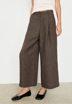 Weit geschnittene braune Leinenhose, getragen mit schwarzen Ballerinas und weißem ärmellosem Top an einem stehenden Model vor einfarbigem Hintergrund.