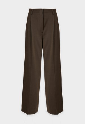 Mujer que lleva una camisa azul clara de botones, pantalones de talle alto marrón oscuro, cinturón rojo y tacones negros puntiagudos con suela roja.