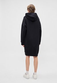 Čierne oversized šaty s kapucňou, žebrovanými manžetami a lemom; majú jednoduchú textúru a neformálnu dĺžku, kombinované s bielymi teniskami.