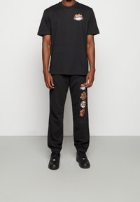 Czarna bawełniana koszulka z kolorowymi logo na piersi, połączona z czarnymi dresami z wielokolorowymi łatami po boku. Czarne sneakersy.