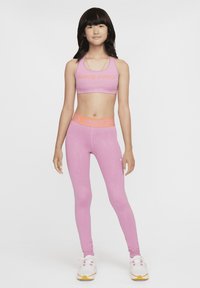 Ružový športový podprsenka Nike Pro a zodpovedajúce legíny s oranžovými akcentmi loga. Materiál vyzerá elasticky; hladká textúra, priliehavý dizajn. Biele tenisky.