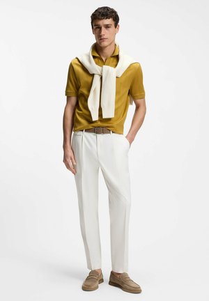 Mann trägt senfgelbes Poloshirt, weißen Pullover über die Schultern gelegt, weiße Hose, braunen Gürtel und beige Loafer, steht mit einer Hand in der Tasche.