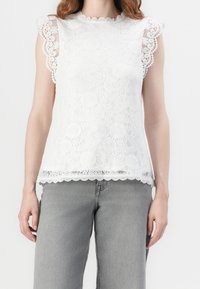 Vit spetsblus med vågiga ärmar och nederkant. Invecklade blommönster genom hela plagget. Kombinerad med ljusgrå vida jeans.