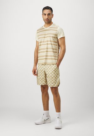 Hombre de pie con las manos en una camiseta de rayas beige, pantalones cortos de cuadros beige, calcetas blancas y zapatillas blancas Nike sobre un fondo liso.