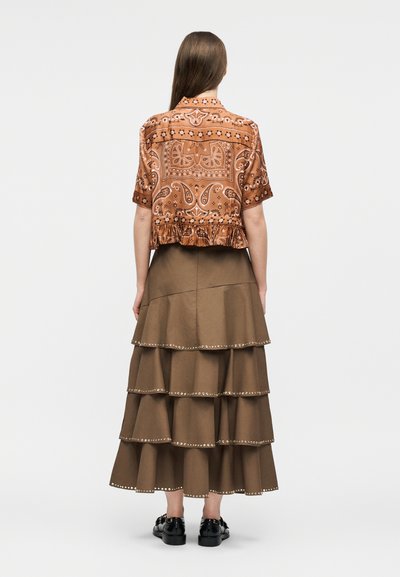 Γυναίκα που φοράει καφέ μπλούζα με μοτίβα και κοντά μανίκια και μακριά πράσινη φούστα με στρώσεις και διακοσμητικές τρουκς στα τελειώματα, στέκεται με την πλάτη προς τα εμπρός.