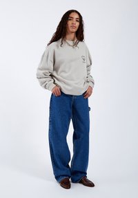 Felpa grigia chiara con grafica a forma di cuore sul petto, abbinata a jeans denim blu larghi. Presenta un colletto rotondo e una vestibilità oversize.