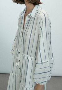 Robe en lin rayée avec de longues manches évasées, devant à boutons, taille avec cordon de serrage, et détail décoratif à pompons en bleu et blanc.