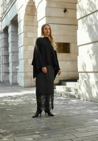 Svart stickad poncho i kombination med en svart kjol med spetsdetaljer. Modellen bär ankelskorts stövlar. Urban miljö med stenarkitektur.