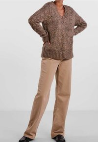 Brauner gestrickter Pullover mit V-Ausschnitt und strukturiertem Muster, kombiniert mit khakifarbenen Hose mit geradem Bein, getragen mit schwarzen Schuhen.