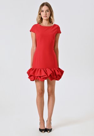 Rotes Kleid mit eng anliegendem Oberteil und kurzen Ärmeln, das einen frillierten Saum hat. Glatte Textur, knielanges Design, kombiniert mit schwarzen Absätzen.
