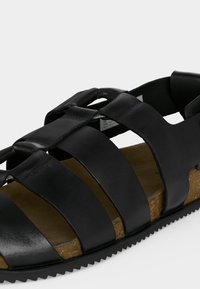 Μαύρο δερμάτινο σανδάλι με πολλαπλούς φαρδιούς ιμάντες, φερμουάρ από φελλό και ραβδωτή μαύρη σόλα σε ανοιχτό φόντο.