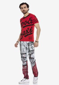 Rotes T-Shirt mit schwarzem Grafiktext; weiße und graue Jeans mit Farbklecksmuster; strukturiertem Stoff; kombiniert mit weißen Sneakern.