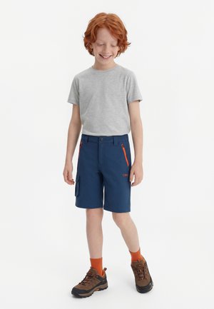 Κοκκινομάλλικο αγόρι που χαμογελά, φορά γκρι μπλουζάκι, σκούρο μπλε σορτς με πορτοκαλί τσέπες με φερμουάρ, πορτοκαλί κάλτσες και καφέ ορειβατικά παπούτσια, στέκεται μπροστά σε άσπρο φόντο.
