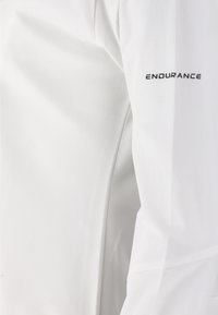 Vit atletisk tröja med en mjuk textur, med en "ENDURANCE"-logga i svart på vänster ärm. Enkel, strömlinjeformad design.
