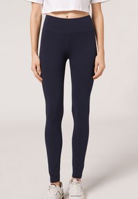 Kvinna som bär marinblå högmidjade leggings, avkortad vit t-shirt och vita sneakers, som står framför en neutral ljus bakgrund.