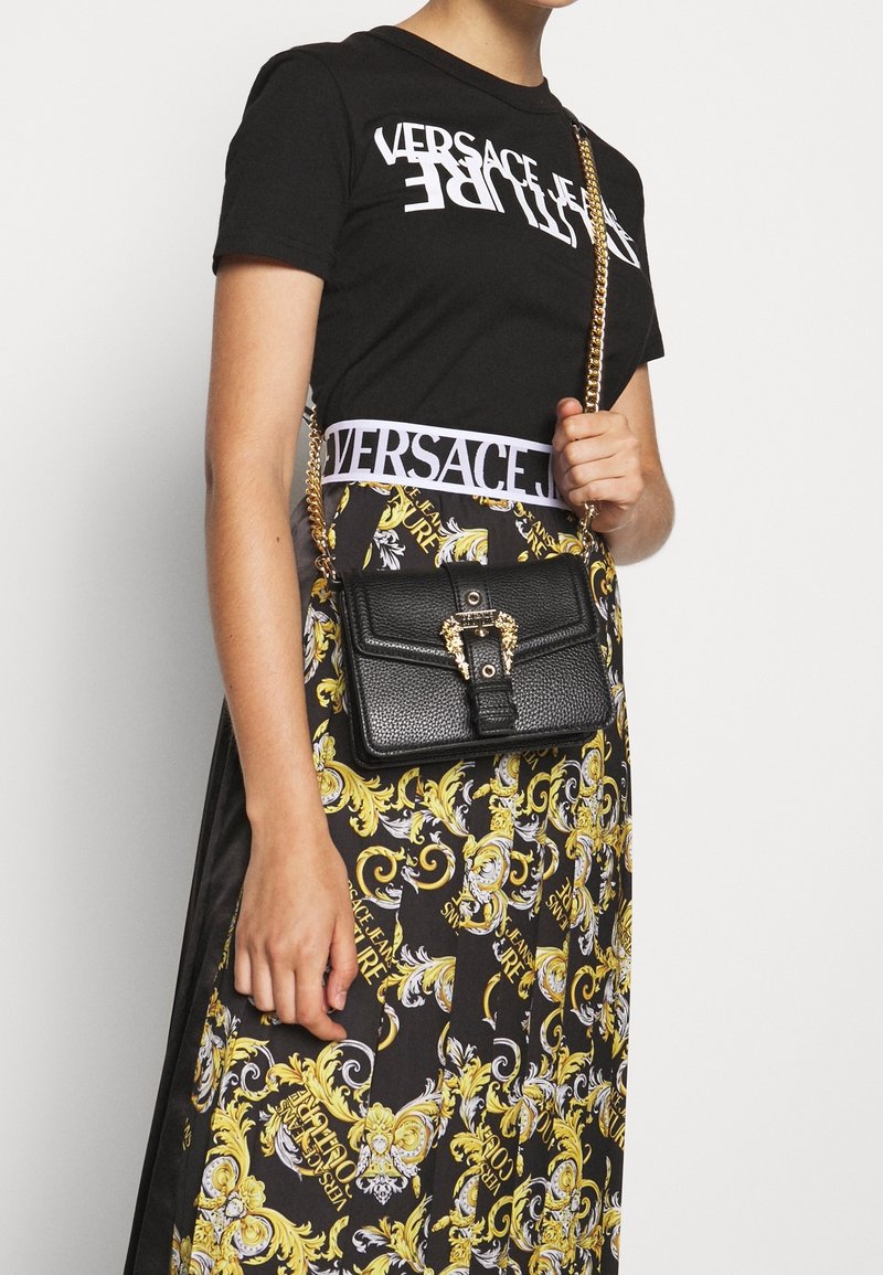 Kvinna iklädd svart Versace t-shirt och mönstrad kjol med gul barockdesign, som håller en svart texturerad handväska med guldkedjestrap.