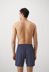 Shorts de bain bleu marine avec une taille élastique, texture lisse et une longueur midi-cuisses, montrés de dos sur un torse nu.