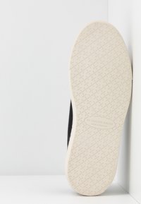 Sneaker nera con suola in gomma bianca e testurizzata. Presenta un logo impresso sulla suola, con un design pulito e minimalista.